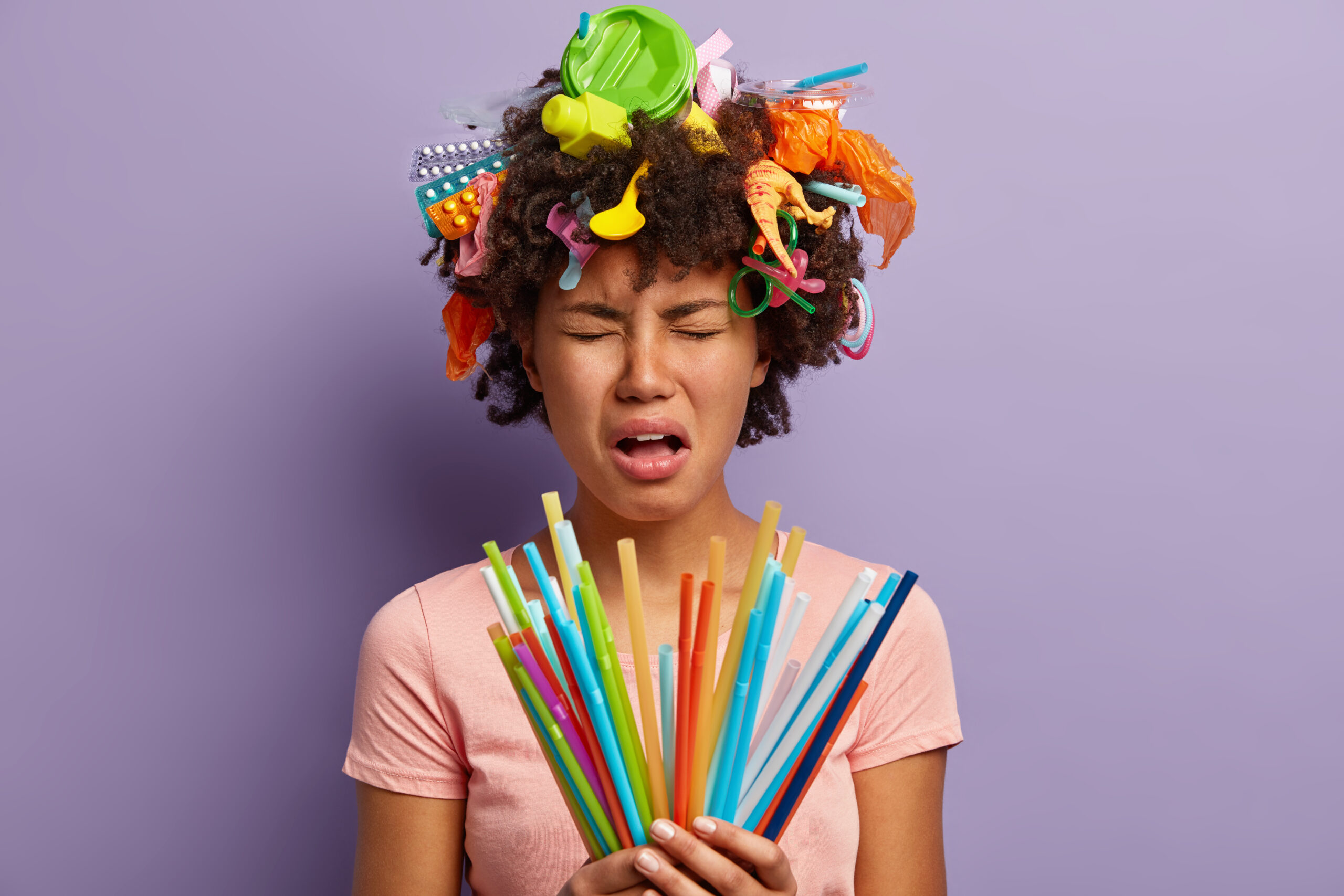 Stop using plastic straws. Depressed young woman with curly hair and dark skin holds bunch of straws, being in panic, begs to save earth, isolated over purple wall. Environmental problem concept