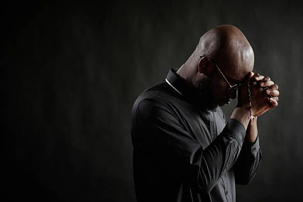 Wide shot of young African American priest with bowed head in black cassock praying silently, he holding praying beads